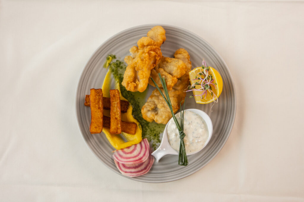 Deep-fried fish, thick chips, a dollop of remoulade, pickled radish slices and a slice of lemon lie on a gray plate. The dish, garnished with herbs and sprouts, stands elegantly on a white tablecloth.