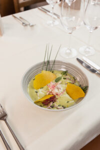 A bowl of gourmet ravioli with a creamy sauce, garnished with crispy orange chips, chives and microgreens. The table is set with a white tablecloth, a fork, a knife and empty wine glasses.