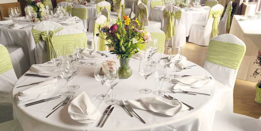 A round table for a formal banquet is set with a white tablecloth, neatly folded napkins, several glasses and utensils, and a colorful flower arrangement in the center; the chairs are adorned with green sashes.