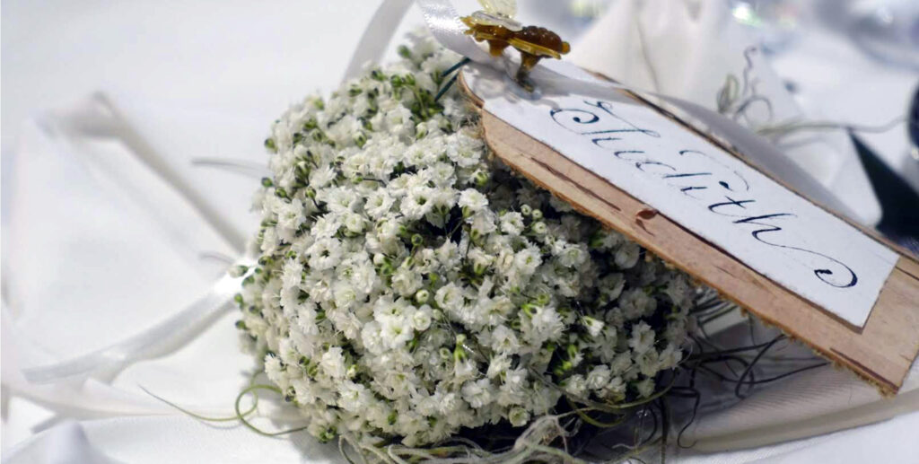 A bouquet of small white flowers with green stems rests on a white cloth, which is adorned with a wooden pendant with elegant calligraphy and the inscription 