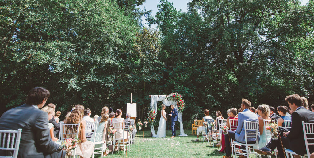 An outdoor wedding ceremony where guests are seated on white banquet chairs facing the bride and groom who stand under a decorated arch surrounded by lush green trees.