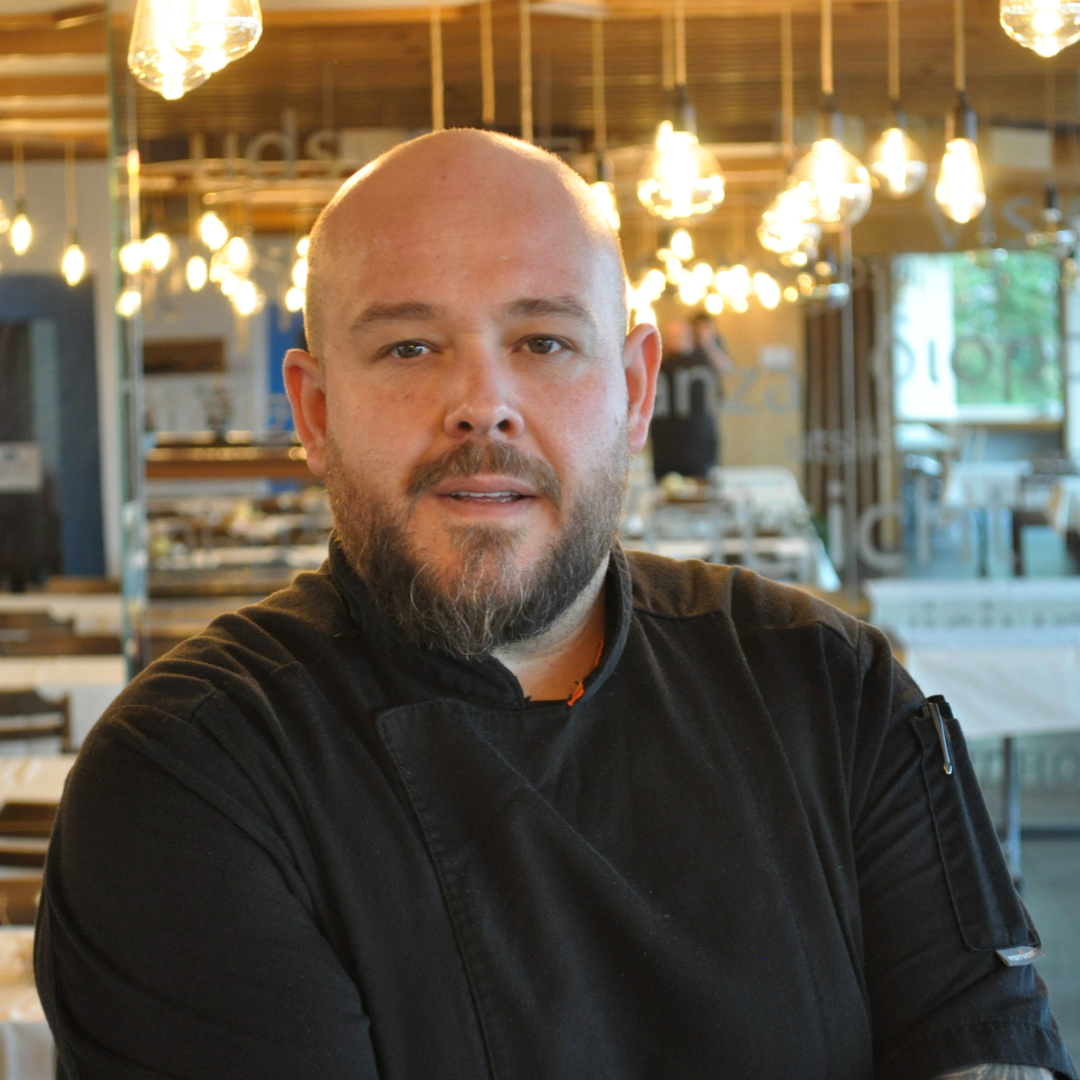 A bearded man in a black chef's jacket stands in a restaurant with warm hanging lamps and laid tables in the background and looks confidently into the camera.