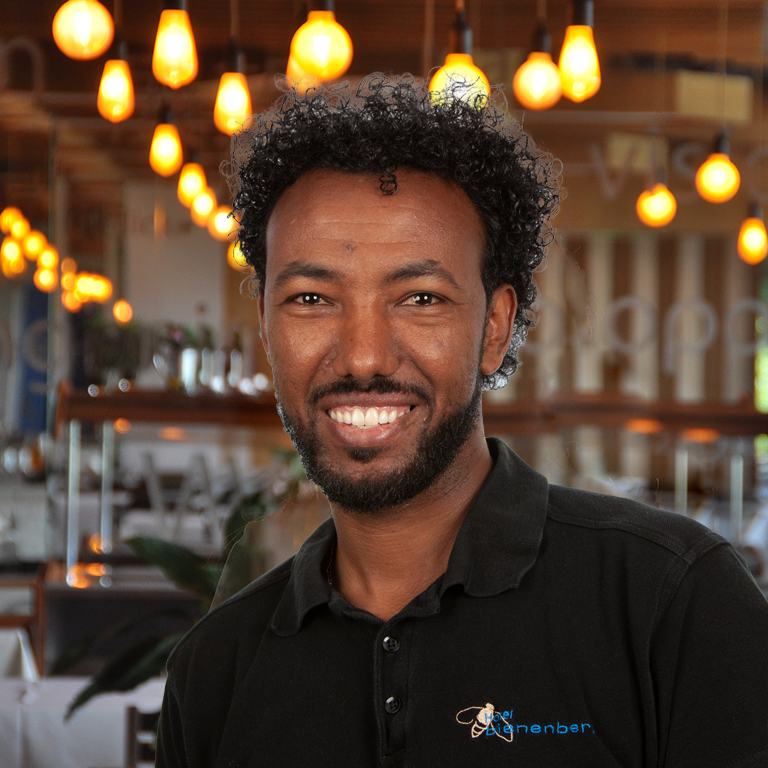 A smiling man with curly hair and a beard stands in a warmly lit restaurant wearing a black shirt with a logo. Hanging light bulbs and blurred tables can be seen in the background.