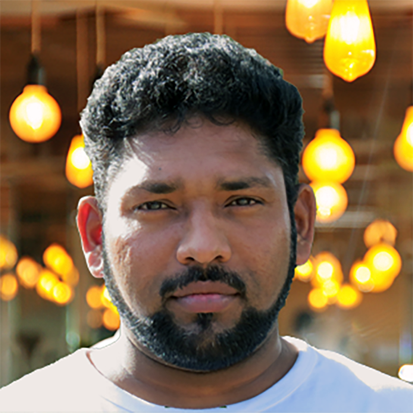 A man with curly dark hair and a beard faces the camera, surrounded by yellow hanging lamps in a warmly lit environment.