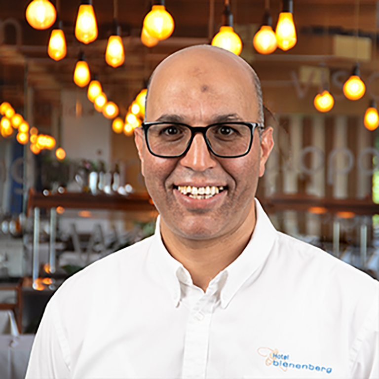 A smiling man with glasses and a white shirt stands in an interior with warm hanging lamps and a blurred restaurant background behind him.