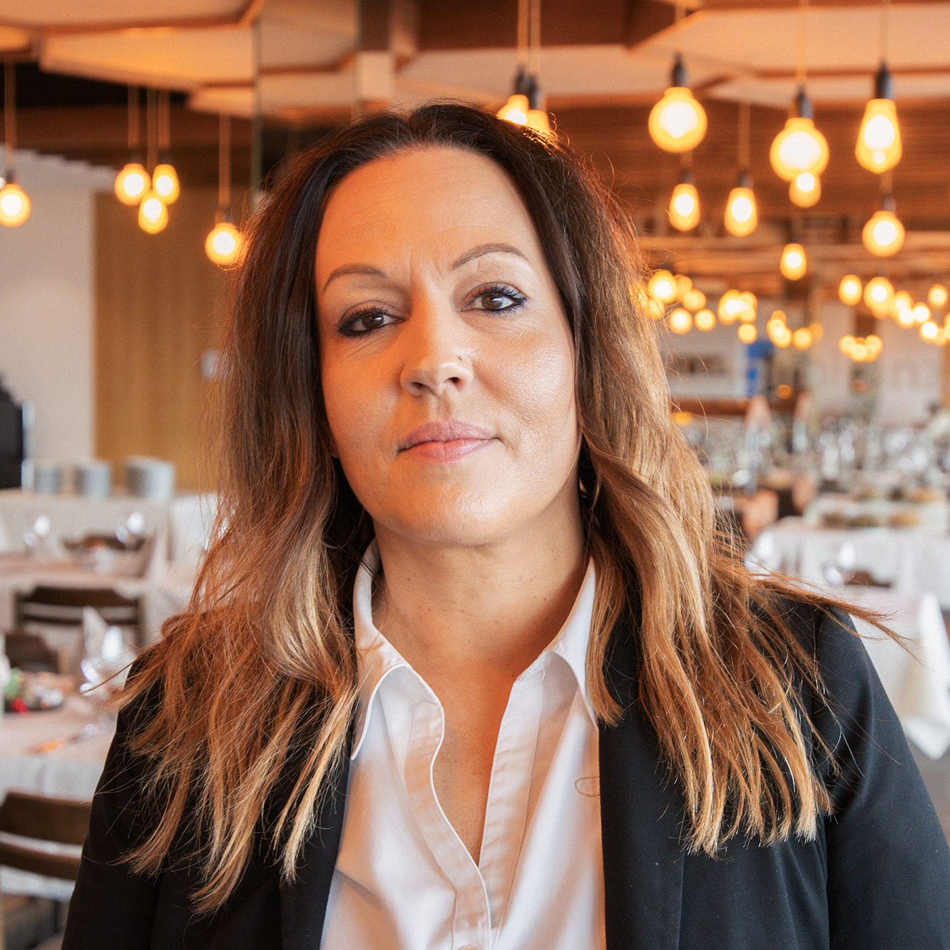 A woman with long, wavy brown hair, wearing a white blouse and a black blazer, stands in a warmly lit restaurant with hanging lamps and neatly laid tables in the background.