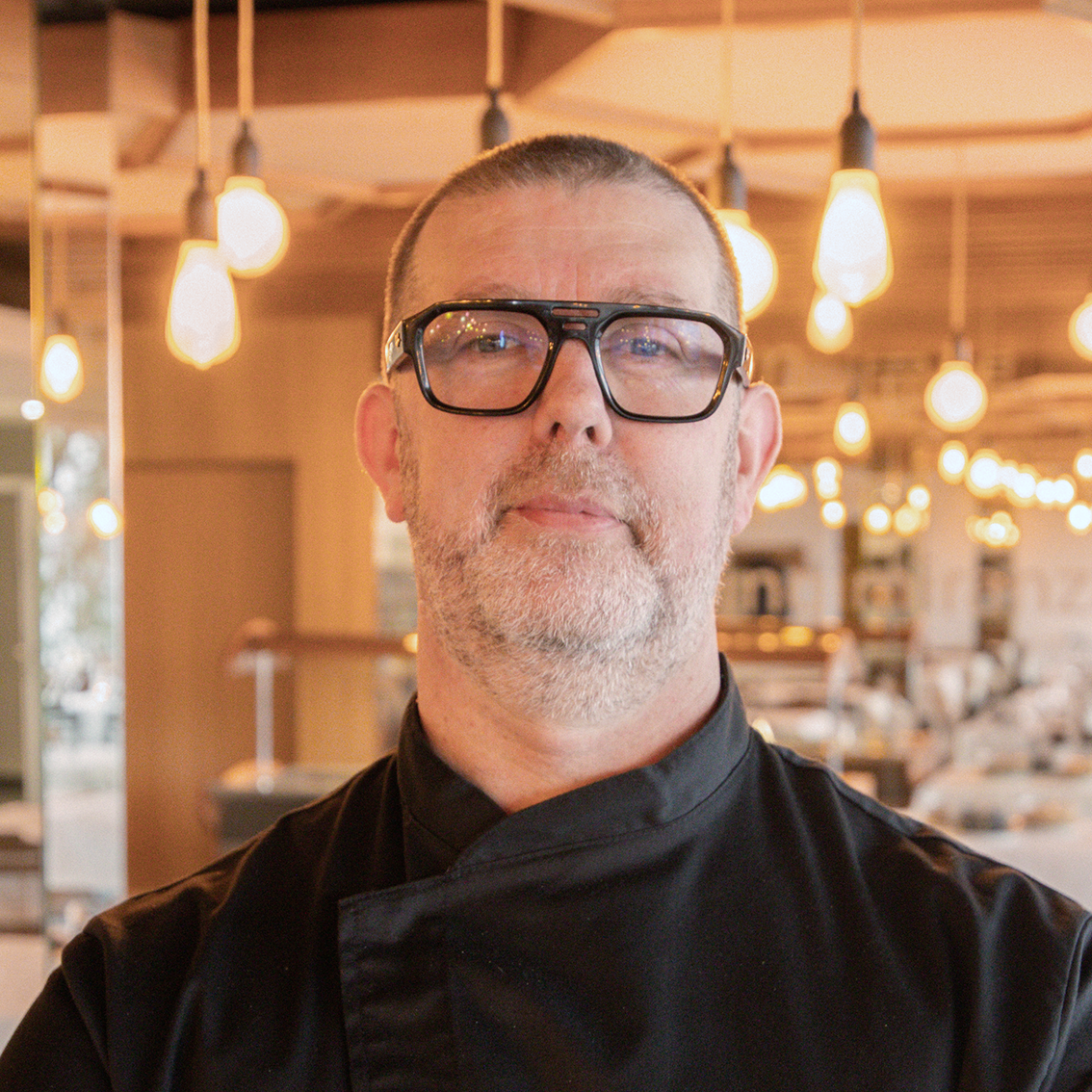 A man with short hair, beard and glasses wears a black chef's jacket and stands in a well-lit restaurant with hanging light bulbs and wooden decor in the background.