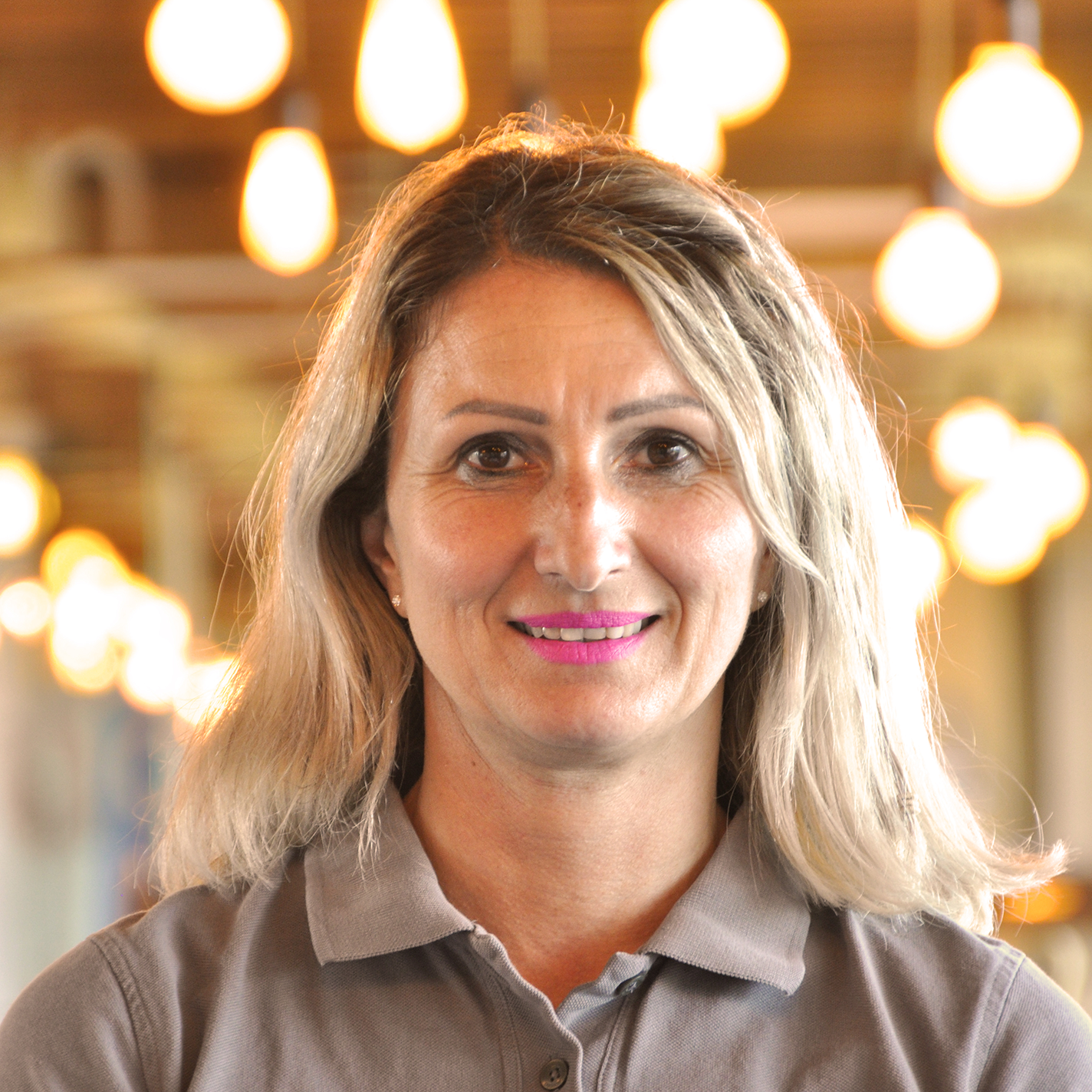 A woman with blonde hair and a gray collared shirt smiles at the camera. Warm lights hang in the blurred background and create a cozy atmosphere.
