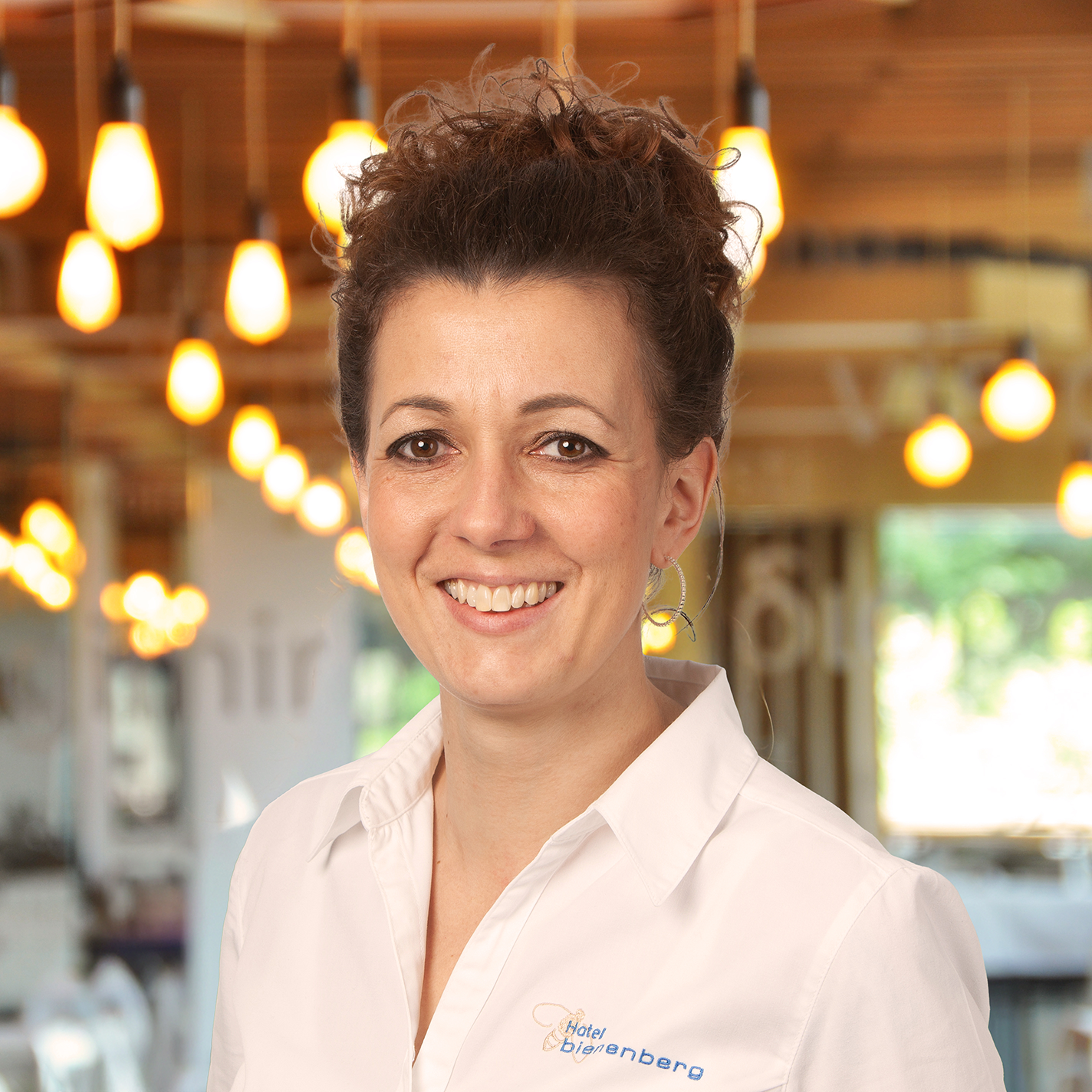 A woman with curly brown hair and a white shirt stands smiling in a warmly lit, modern interior with hanging lamps and a blurred background.