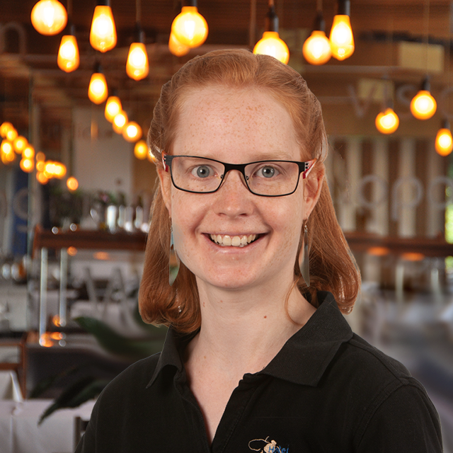 A smiling person with red hair, glasses and a black collared shirt stands in an interior with warm hanging lamps and a blurred restaurant background.