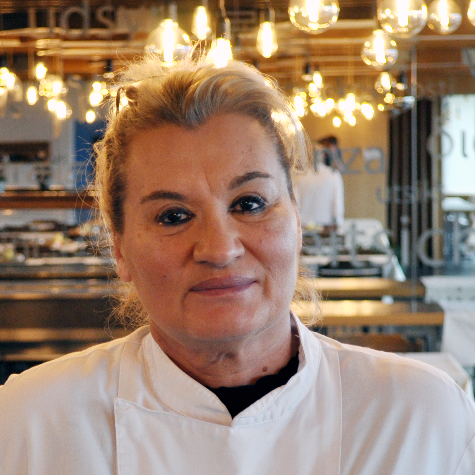 A woman with blonde hair wearing a white chef's coat stands in a modern restaurant with hanging lamps and blurred background elements.