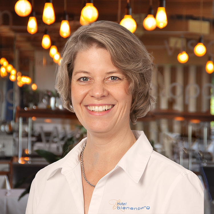 A smiling woman with short, light brown hair stands in a room wearing a white shirt with the words 