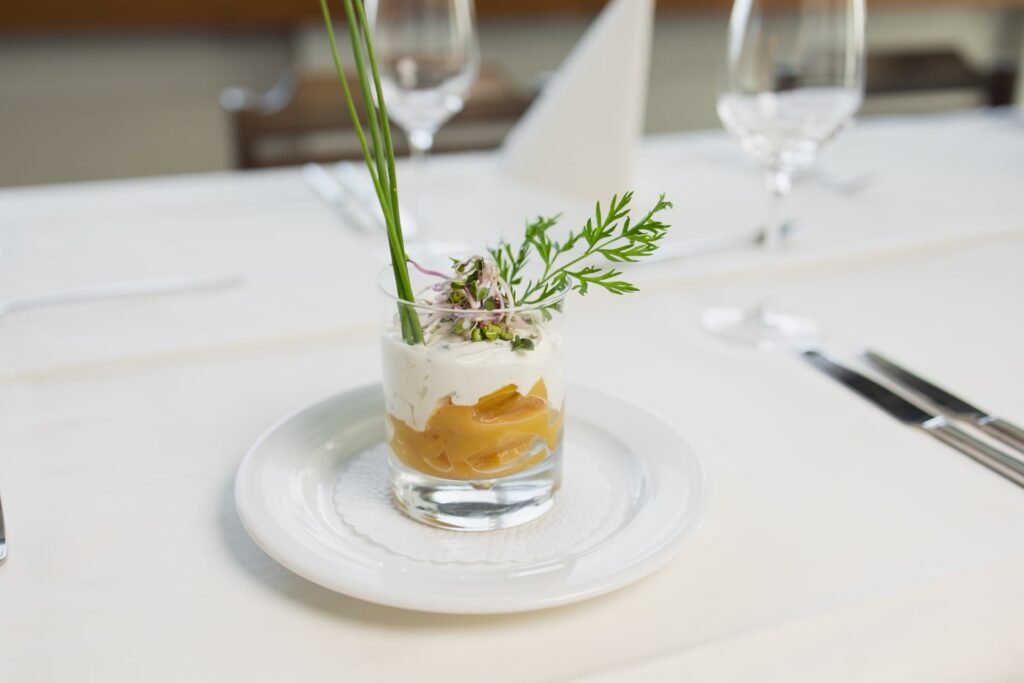 Ein Glasbecher mit einem Dessert oder einer Vorspeise mit gelber Fruchtschicht und weißer Sahnesauce, garniert mit Kräutern, steht auf einem weißen Teller - perfekt für eine elegante Hochzeitstafel.