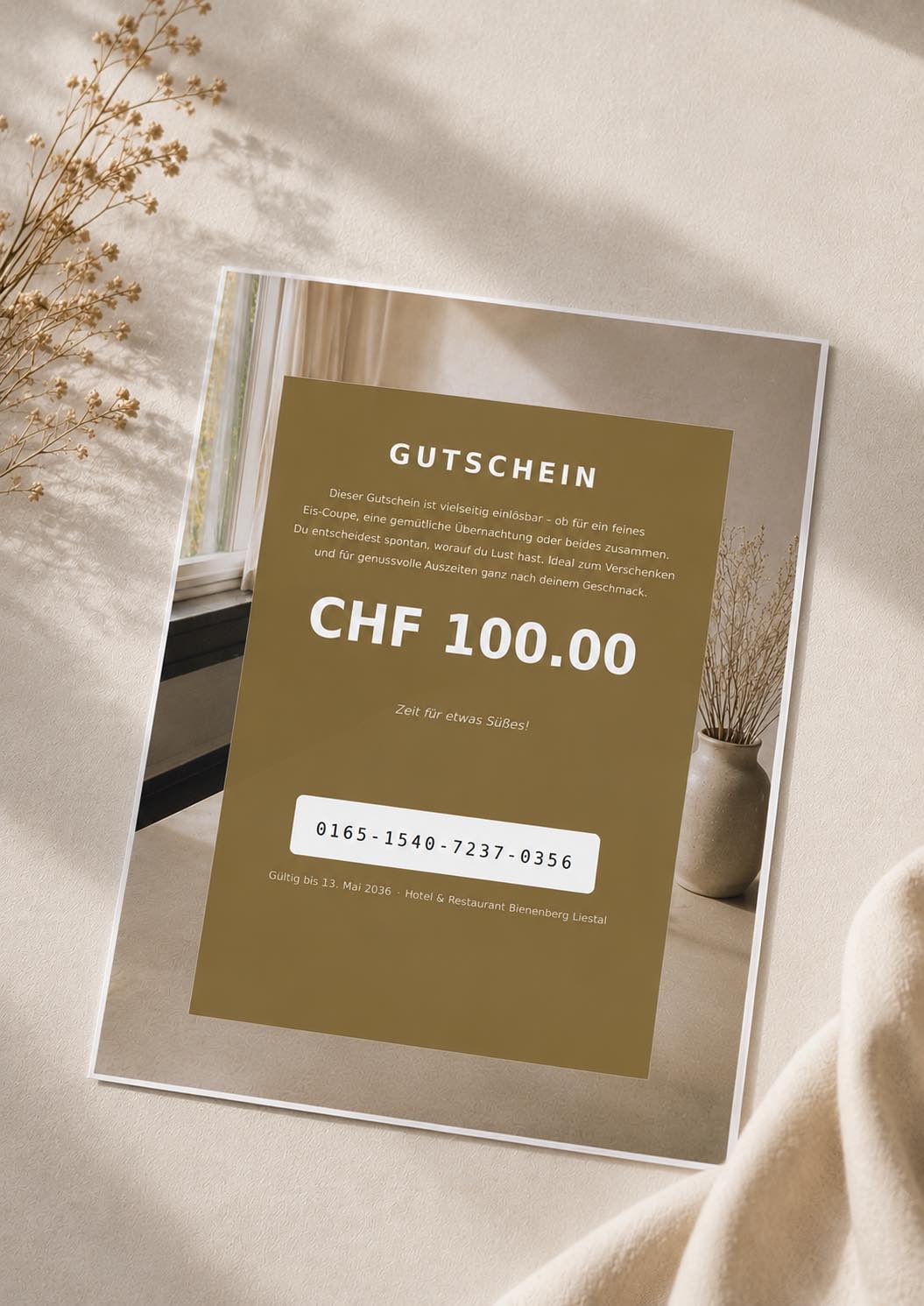 Ein Geschenkgutschein im Wert von CHF 100 liegt auf einer beigen Fläche mit Trockenblumen daneben. Es handelt sich um einen Hotel- und Restaurantgutschein mit Code und Gültigkeitsdatum, der in einem modernen, minimalistischen Design gehalten ist.