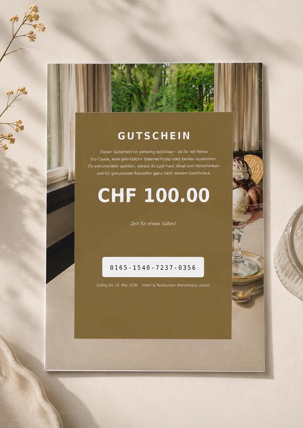 Ein Gutschein im Wert von "CHF 100.00" liegt auf einem Tisch, umgeben von Dessertschalen und Blick ins grüne Gartensetting. Dekorative Trockenblumen und ein Glasornament runden den Gutschein Coup stilvoll ab.