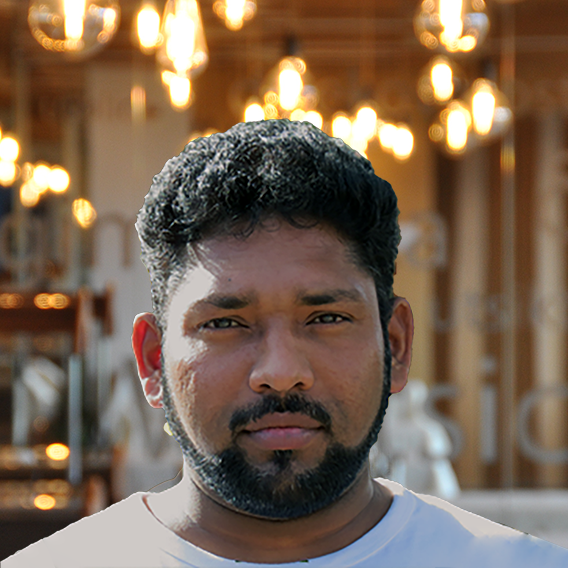 A man with short, curly hair and a beard stands in front of a background decorated with hanging, glowing lightbulbs. He is looking directly at the camera with a neutral expression.