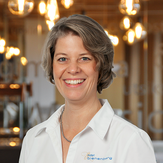 A smiling woman with short blonde hair and a white collared shirt stands in an indoor space with warm lighting and blurred hanging lamps in the background.