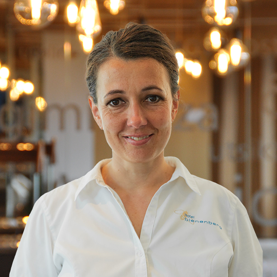 A woman with her brown hair pulled up and a white collared shirt stands indoors in front of warm, blurred lights and glass. She is smiling gently at the camera.